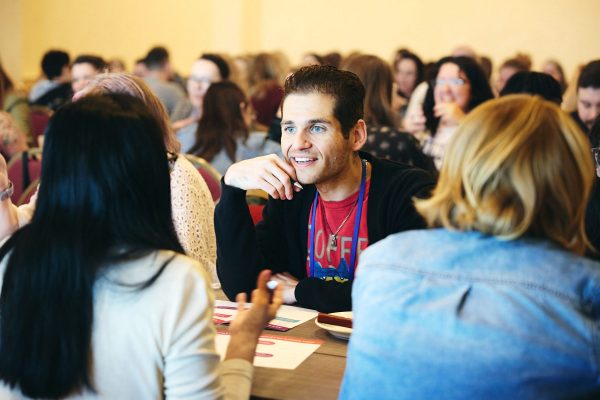 A group of people engage in conversation around a table at a busy event, fostering a culture of kindness and inclusivity.