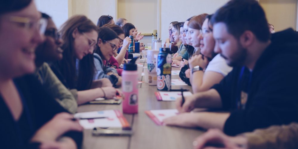 People are seated on both sides of a long table, engaging in conversation and taking notes during a workshop or meeting, fostering a collaborative culture.