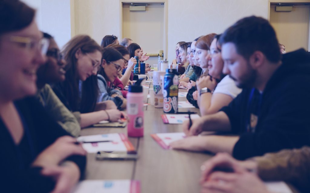 People are seated on both sides of a long table, engaging in conversation and taking notes during a workshop or meeting, fostering a collaborative culture.