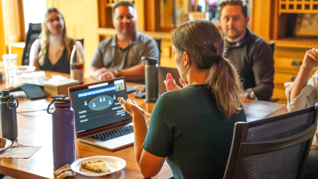A team gathers around a table during a workplace meeting, with one woman presenting insights about effective change management from her laptop screen. The atmosphere is collaborative and engaged.